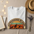 White t-shirt with "OUTNUMBERED BY PAWS" and mountain sunset graphic.