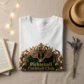 White t-shirt with "Pickleball Cocktail Club" text and ornate gold pickleball paddles.