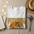 White t-shirt with "PICKLEBALL" graphic of woman playing sport.