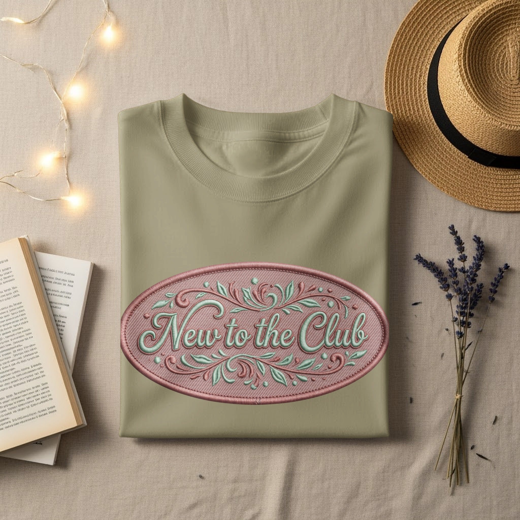 Embroidered "New to the Club" on a light olive t-shirt, with straw hat and lavender.