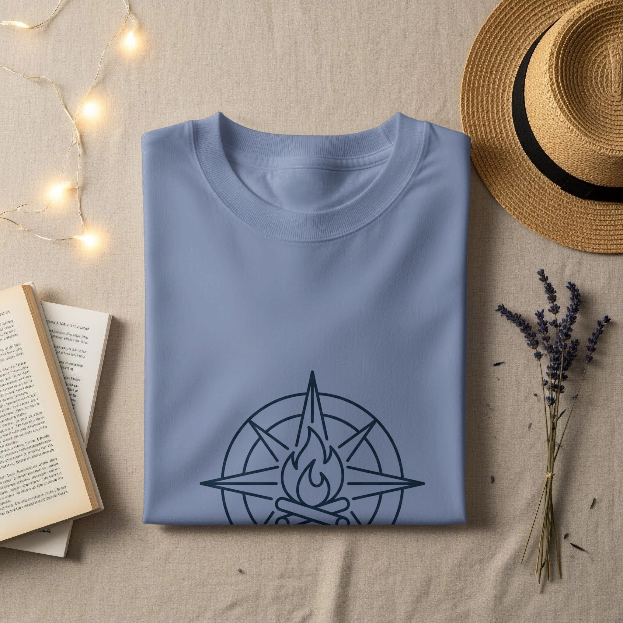 Blue t-shirt with compass and campfire graphic, laid flat with hat and book.