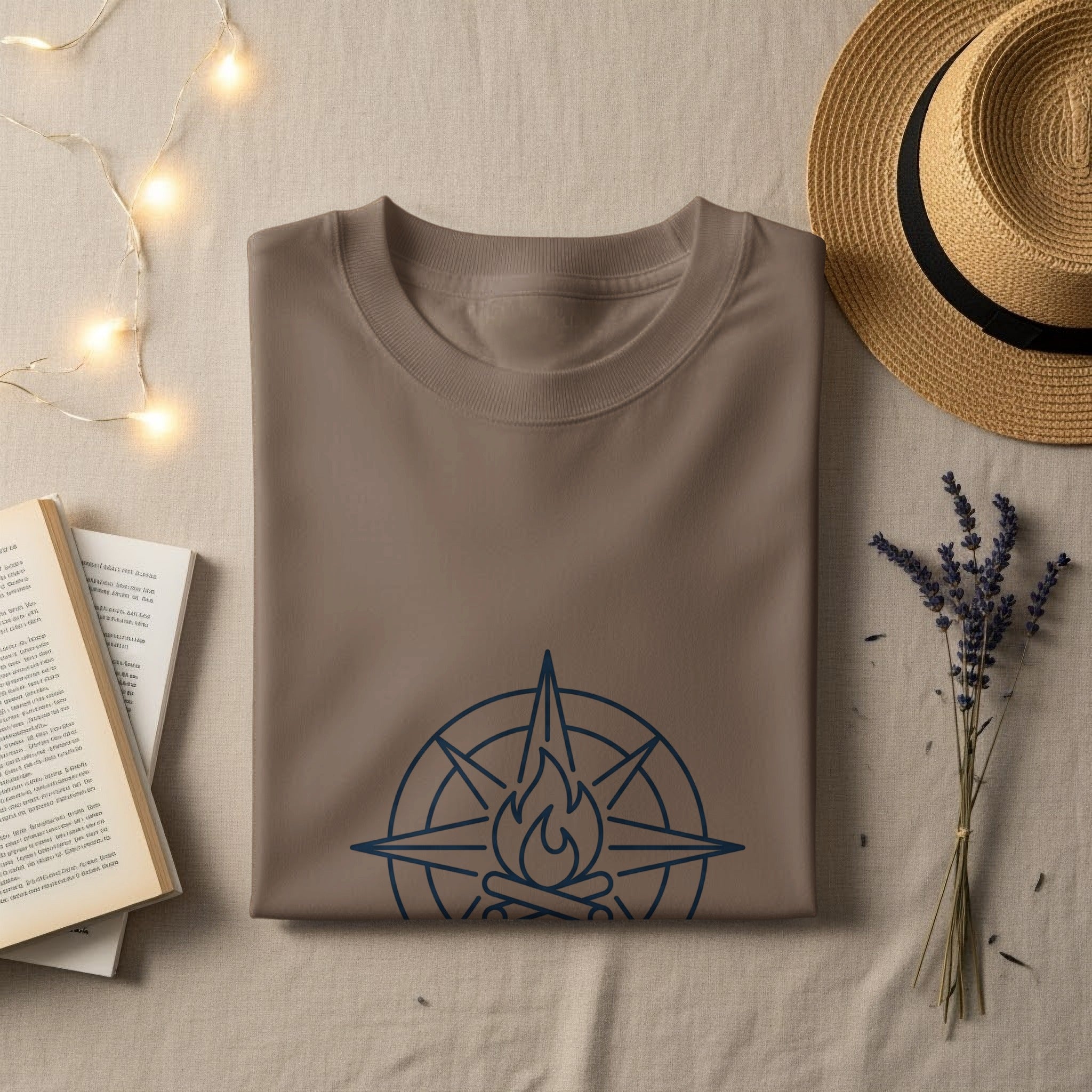 Brown t-shirt with compass and campfire graphic, laid flat with hat and book.