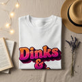 White t-shirt with "Dinks &" in colorful, 3D text.