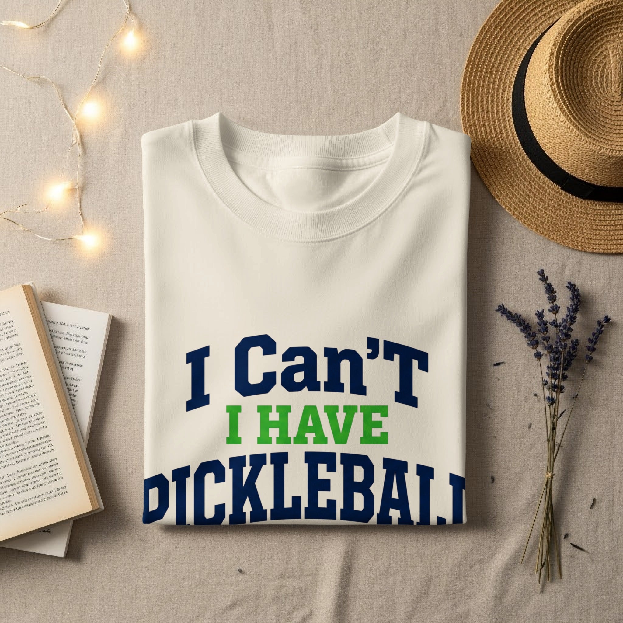 White t-shirt with "I Can't I HAVE PICKLEBALL" text, laid flat with straw hat and books.