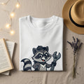 White t-shirt with cartoon raccoon holding a wrench, flat lay with hat and book.